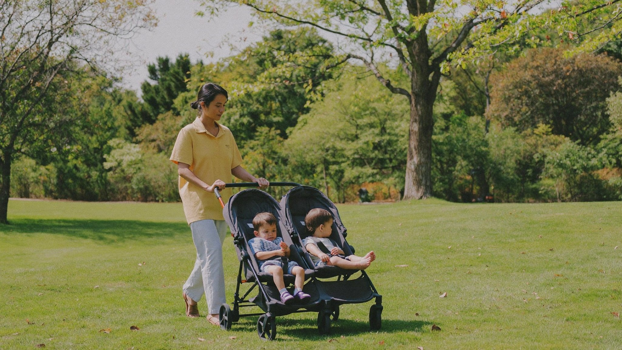Mother with a twin double pram in a leafy park - Bambinos & Beyond Double & Twin Travel Systems